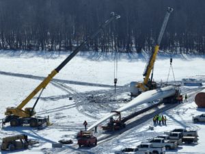 Cranes lifting wind turbine blade on snowy construction site.